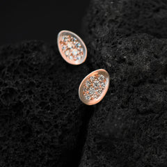 Thin, concave, egg-shaped studs that are pavé set with 10 tiny white diamonds each that follow the egg shape; shown in a crevice between two lava rocks